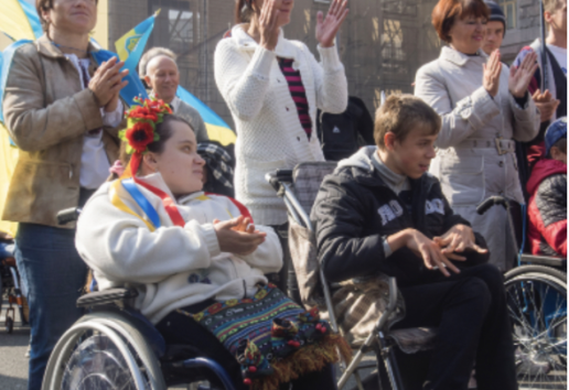 Des personnes en fauteuil roulant participent à une manifestation, entourées de spectateurs applaudissant, tenant des drape