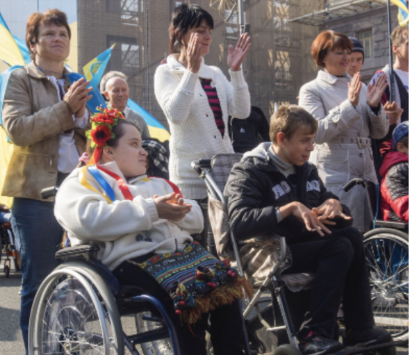Des personnes en fauteuil roulant participent à une manifestation, entourées de spectateurs applaudissant, tenant des drape