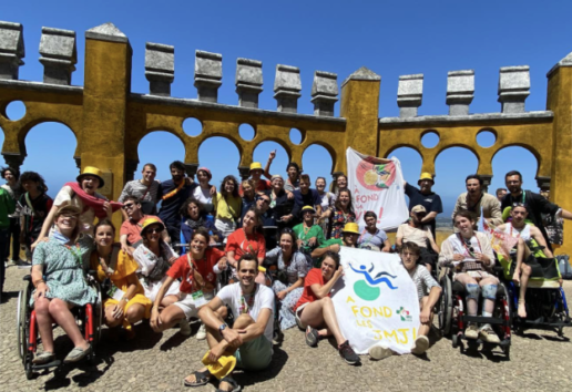 Groupe joyeux devant arcades historiques, incluant personnes en fauteuil roulant, sous un ciel bleu. Événement festif avec
