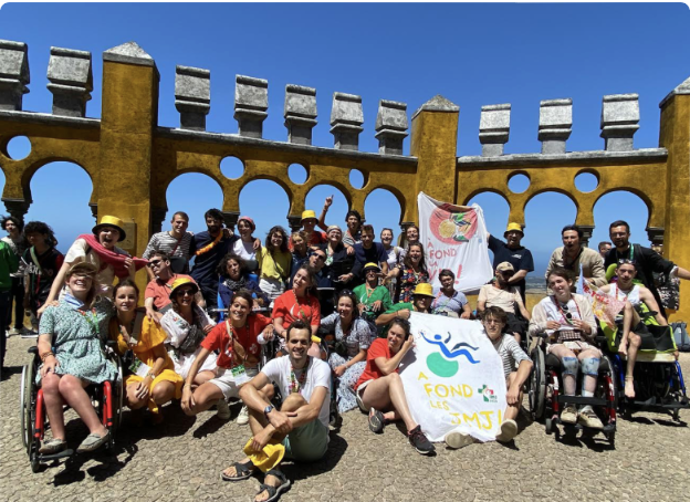Groupe joyeux devant arcades historiques, incluant personnes en fauteuil roulant, sous un ciel bleu. Événement festif avec
