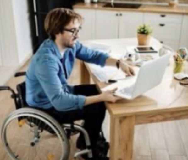Un homme en fauteuil roulant travaille sur un ordinateur portable dans une cuisine moderne, accompagné d'un petit chien à s