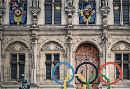 Bâtiment historique avec statues et drapeaux, devant lequel sont superposés les anneaux olympiques colorés.
