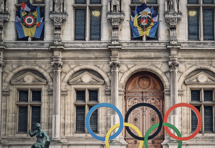 Bâtiment historique avec statues et drapeaux, devant lequel sont superposés les anneaux olympiques colorés.