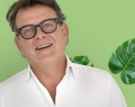 Homme souriant portant des lunettes, vêtu d'une chemise blanche, devant un fond vert avec une feuille de plante.