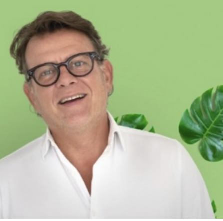 Homme souriant portant des lunettes, vêtu d'une chemise blanche, devant un fond vert avec une feuille de plante.