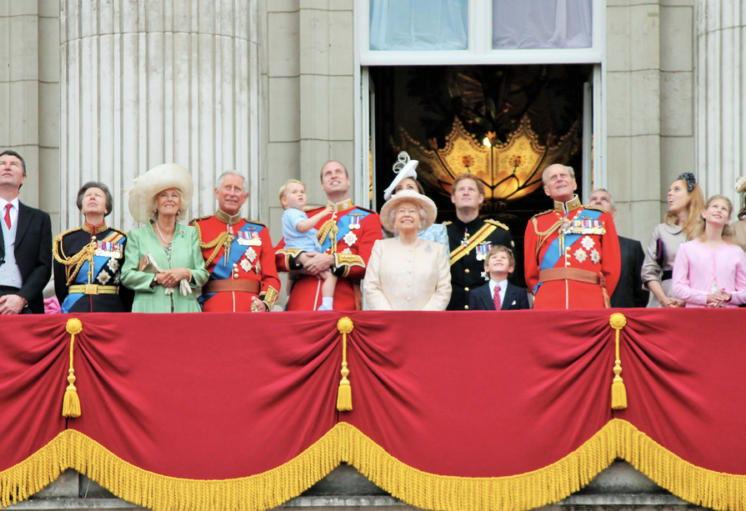 Balcon royal avec membres de la famille royale britannique et invités en tenue officielle, devant un rideau rouge orné.