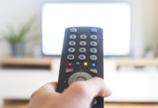Main tenant une télécommande noire devant un écran de télévision allumé.