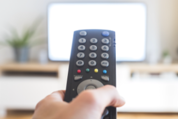 Main tenant une télécommande noire devant un écran de télévision allumé.