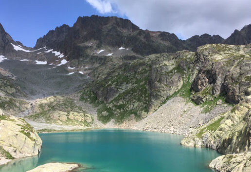Lac alpestre entouré de montagnes rocheuses et de végétation, avec des zones enneigées et un ciel dégagé.