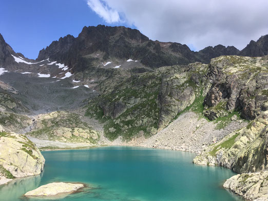 Lac alpestre entouré de montagnes rocheuses et de végétation, avec des zones enneigées et un ciel dégagé.