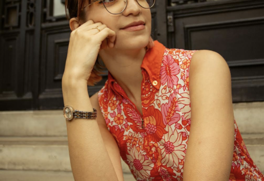Une personne assise pose contre un portail métallique, vêtue d'une chemise à motifs floraux rouge et rose, avec des lunett