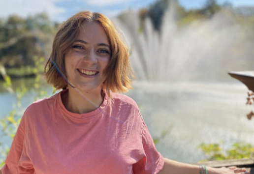 Une personne souriante en rose, debout près d'une balustrade, devant une fontaine et un paysage verdoyant sous un ciel déga