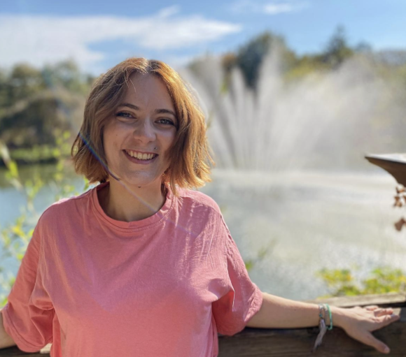 Une personne souriante en rose, debout près d'une balustrade, devant une fontaine et un paysage verdoyant sous un ciel déga