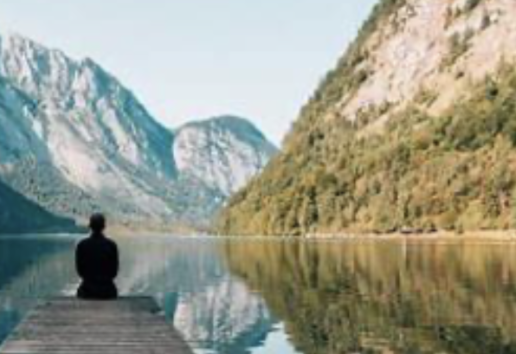Personne assise en méditation sur un pont au bord d'un lac paisible, entouré de montagnes enneigées et de forêts.