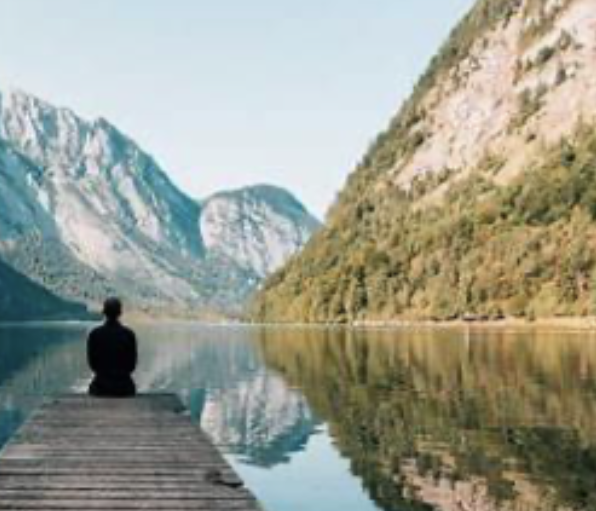 Personne assise en méditation sur un pont au bord d'un lac paisible, entouré de montagnes enneigées et de forêts.