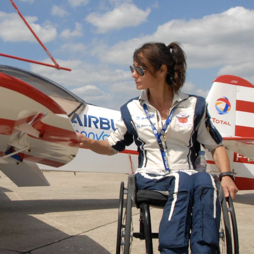 Une personne en fauteuil roulant, vêtue d'un uniforme bleu et blanc avec des logos, pose près d'un avion.