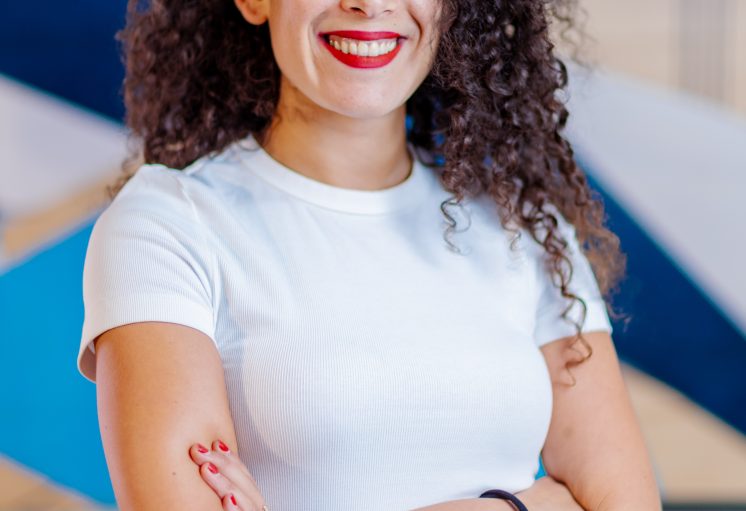 Femme souriante aux cheveux bouclés croisés les bras vêtue d’un t-shirt blanc et d’un jean bleu